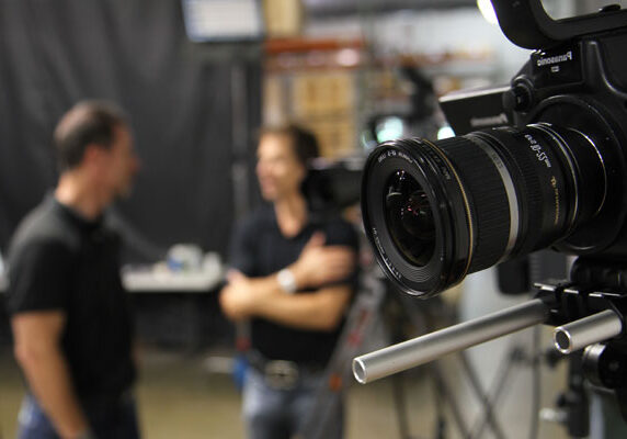 A photo of a video camera with two people in conversation behind the scenes